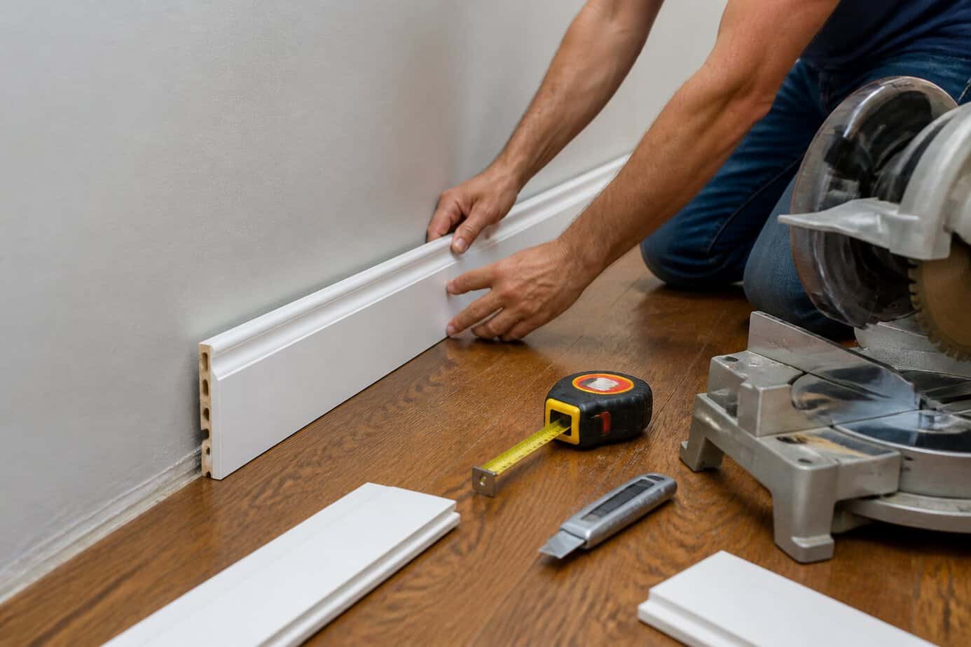 Hands installing new white composite trim along the base of a wall in a bright room with hardwood floors.