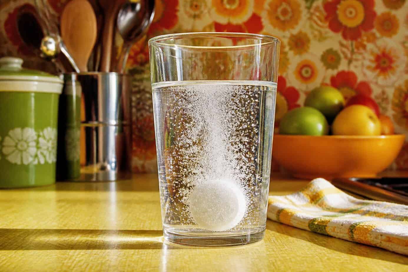 A close-up of a glass of fizzy water with a tablet dissolving inside, surrounded by a retro kitchen setting.