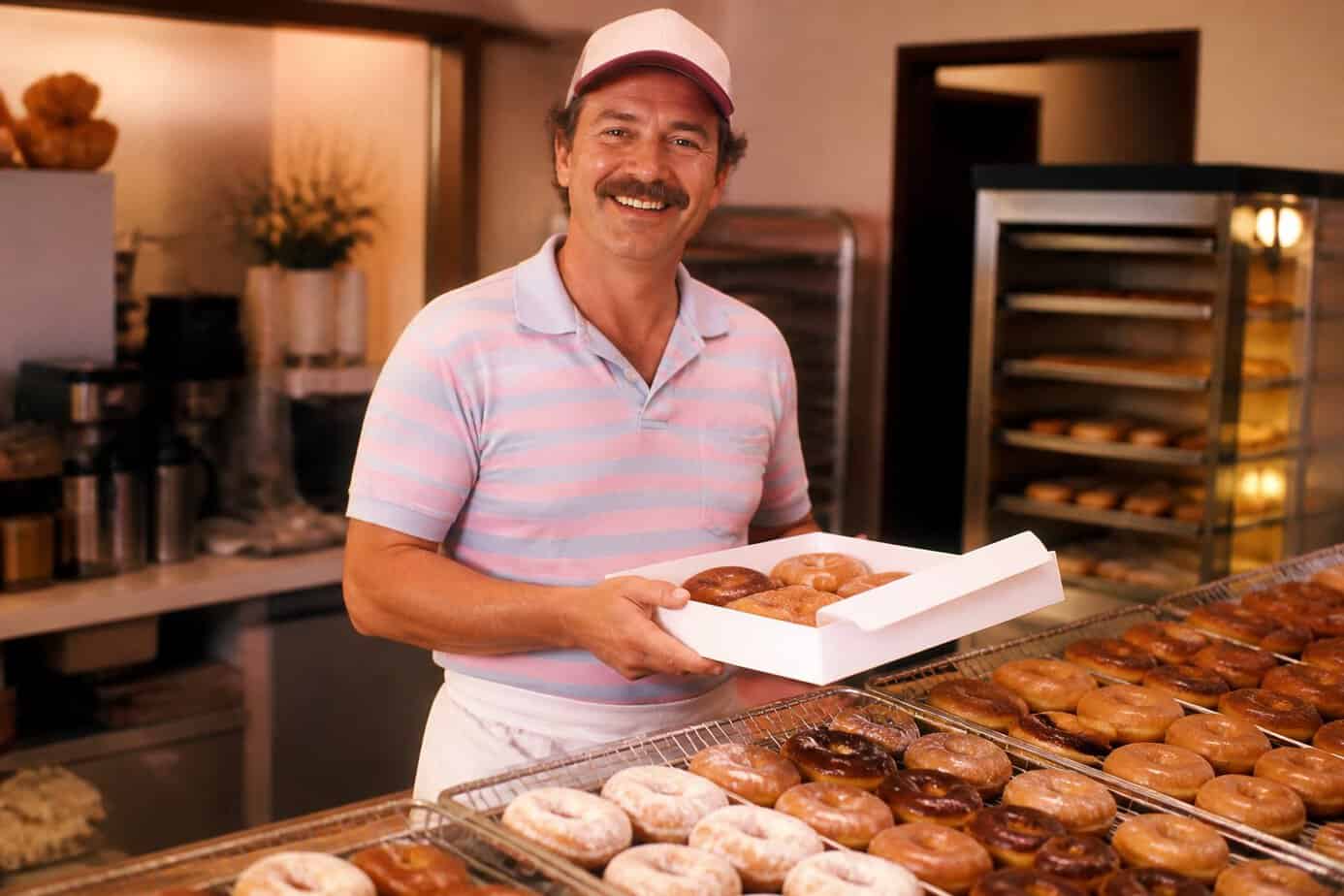 A smiling man in 1980s clothing stands behind a bakery counter filled with fresh donuts in a warm, inviting bakery.