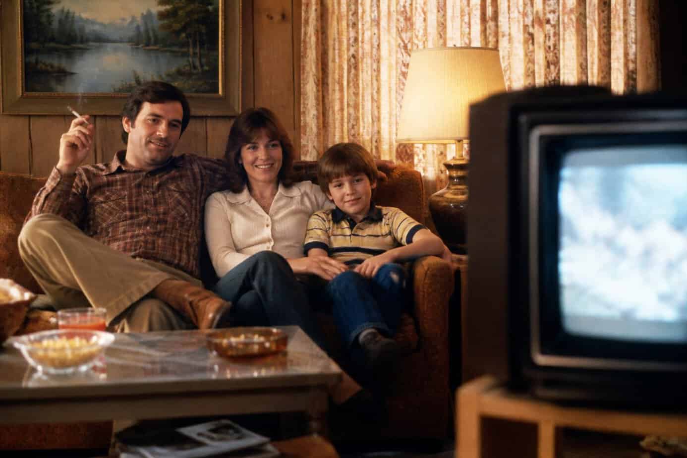A family of three sitting in a vintage living room with a retro television, the father holding a cigarette and everyone looking relaxed and comfortable.