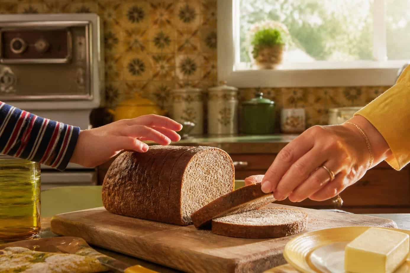 A vintage kitchen scene with a loaf of rye bread on a cutting board and hands reaching for it.