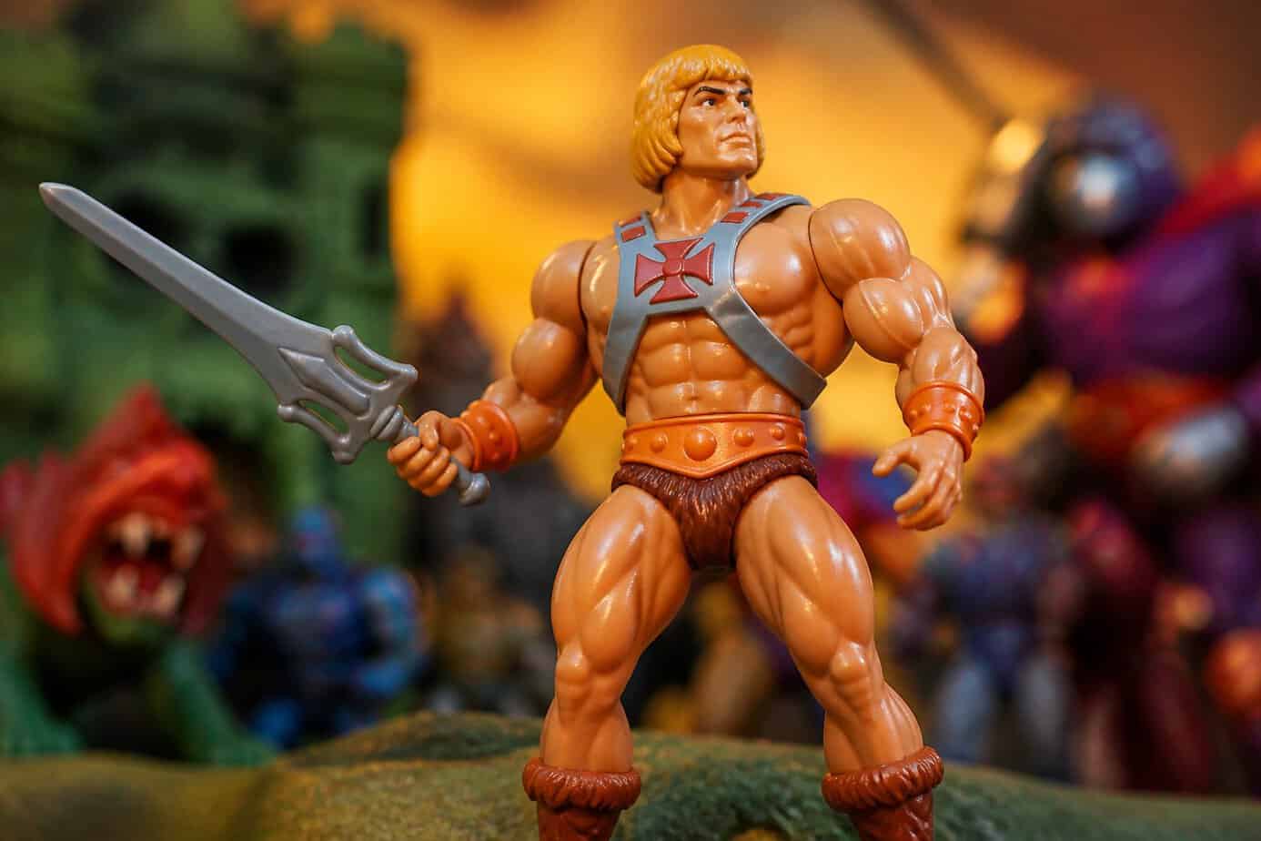 A He-Man action figure posed heroically with a sword, surrounded by blurred 1980s toys in the background.