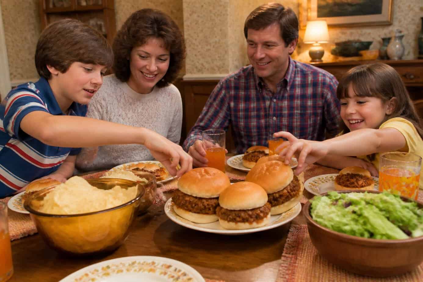 A family gathered around a dining table enjoying sloppy joes and side dishes in a cozy home setting.