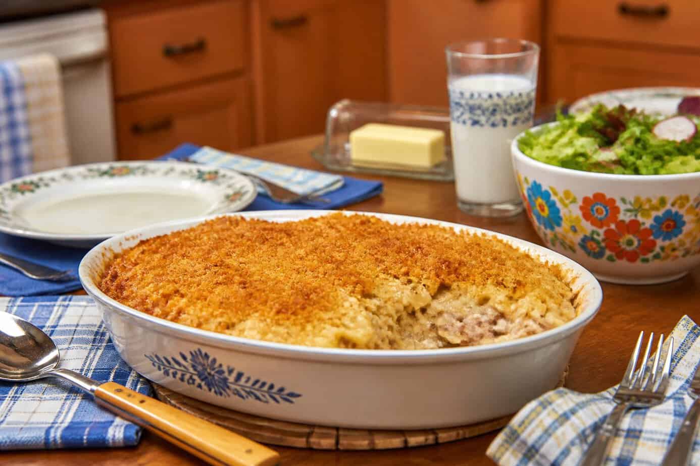 A golden tuna noodle casserole in a ceramic dish on a wooden table with a glass of milk and a salad bowl in a cozy kitchen setting.