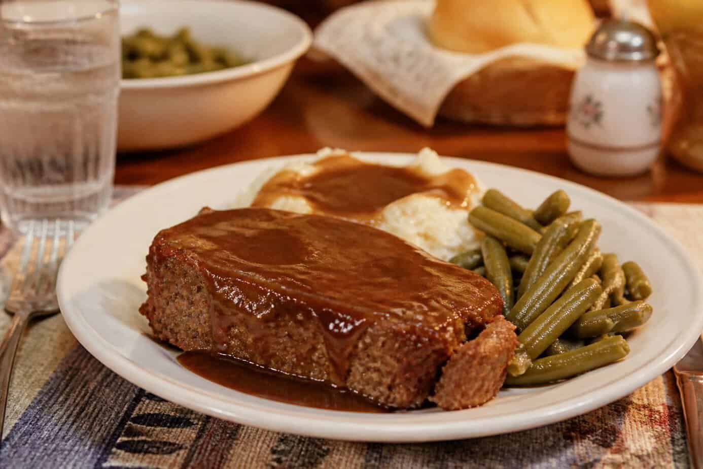 A plate of meatloaf with brown gravy, mashed potatoes, and green beans on a wooden dining table.