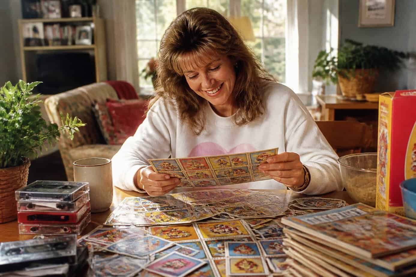 A middle-aged woman sorting colorful trading sticker sheets and collectible items on a wooden table in a sunlit kitchen.