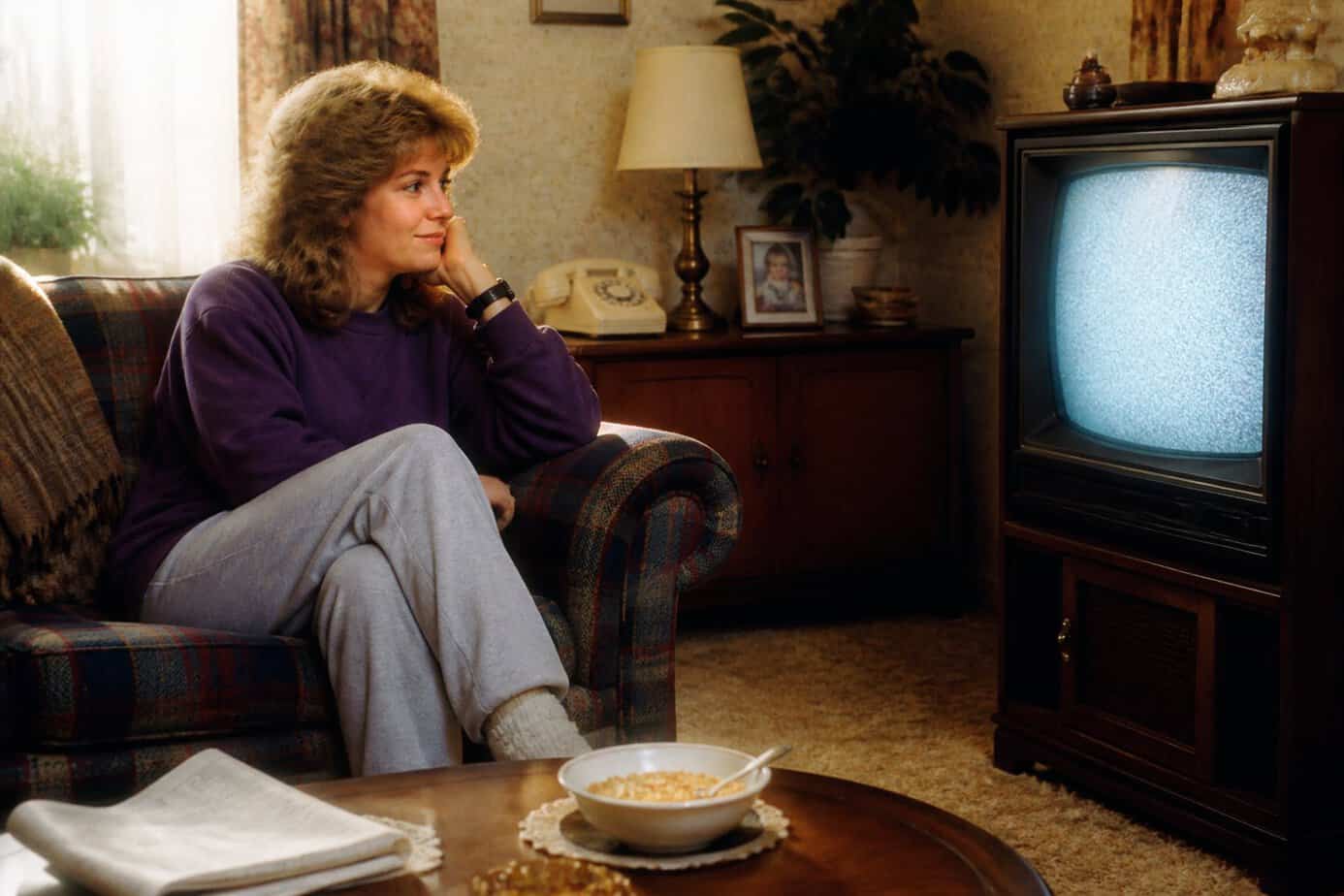 A woman sitting on a couch in a living room, listening to the static from an old television as it warms up.