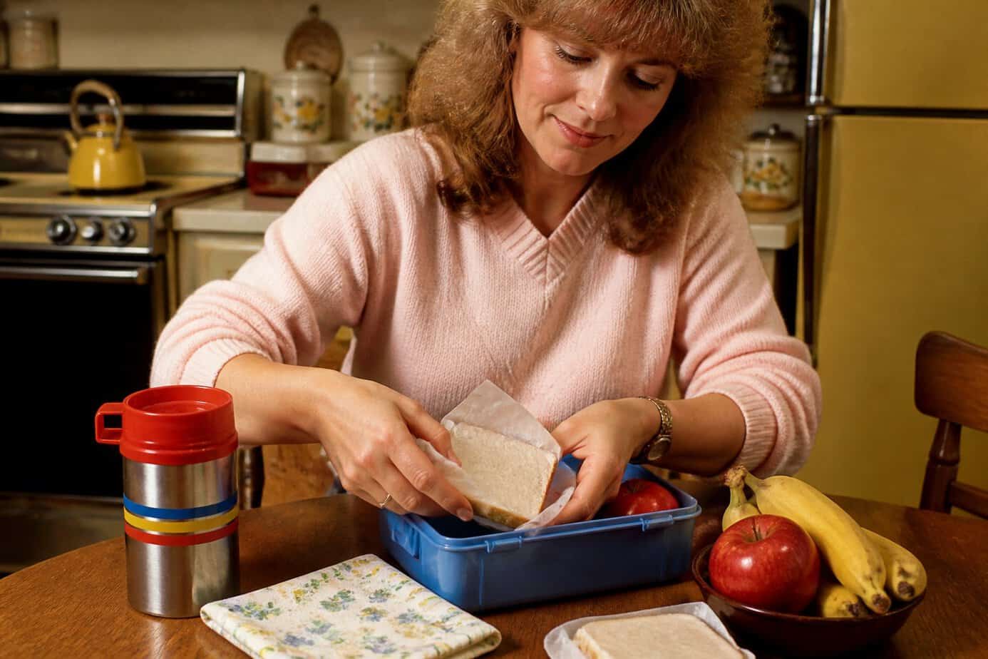 A mother packing a school lunch with a thermos and napkin on a kitchen table.