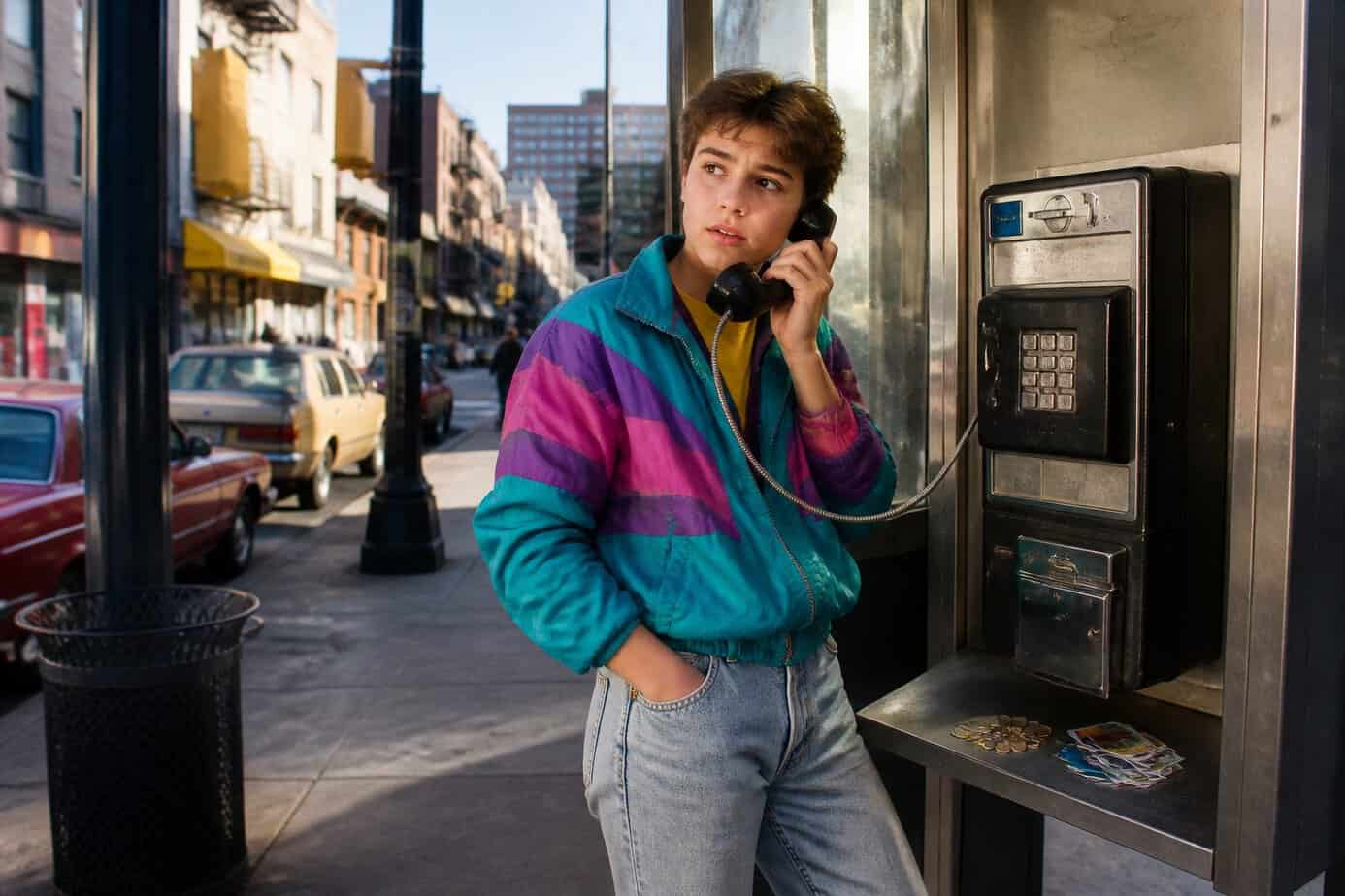 A young person in 1980s clothes using a payphone inside a phone booth on a city street with vintage cars and storefronts in the background.