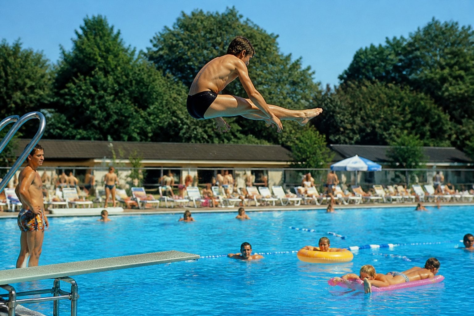 A young person performing a diving board trick at a busy outdoor municipal swimming pool on a sunny day.