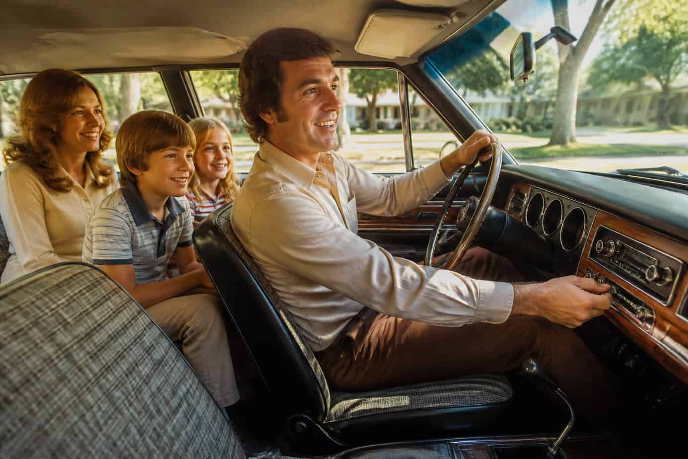A family of four inside a 1970s car listening to the radio while driving through a sunny neighborhood.