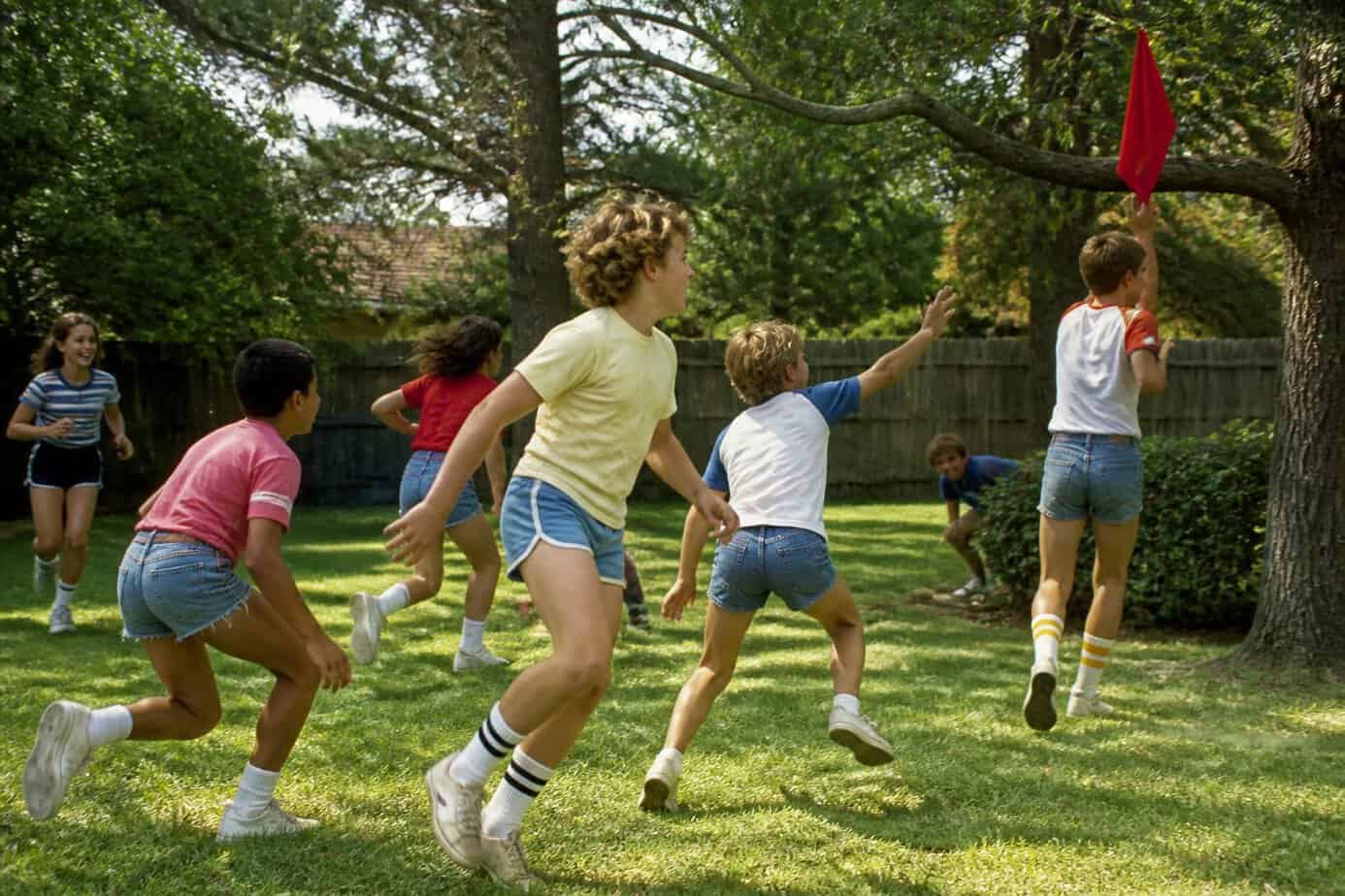 Children playing capture the flag in a sunny backyard surrounded by trees and grass.
