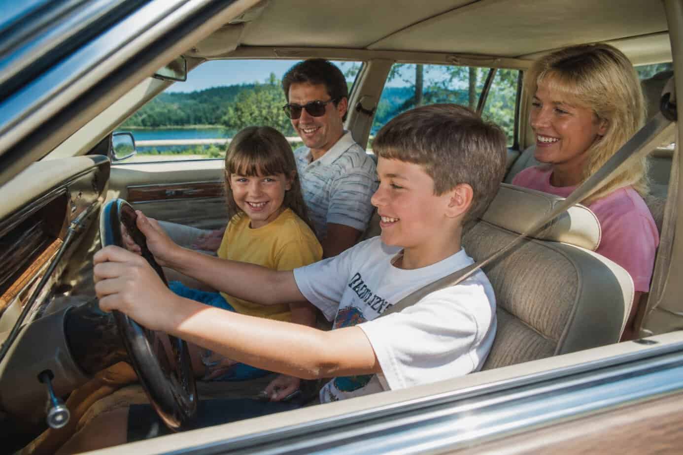 A family in a 1980s station wagon with children pretending to drive while parents watch and smile.