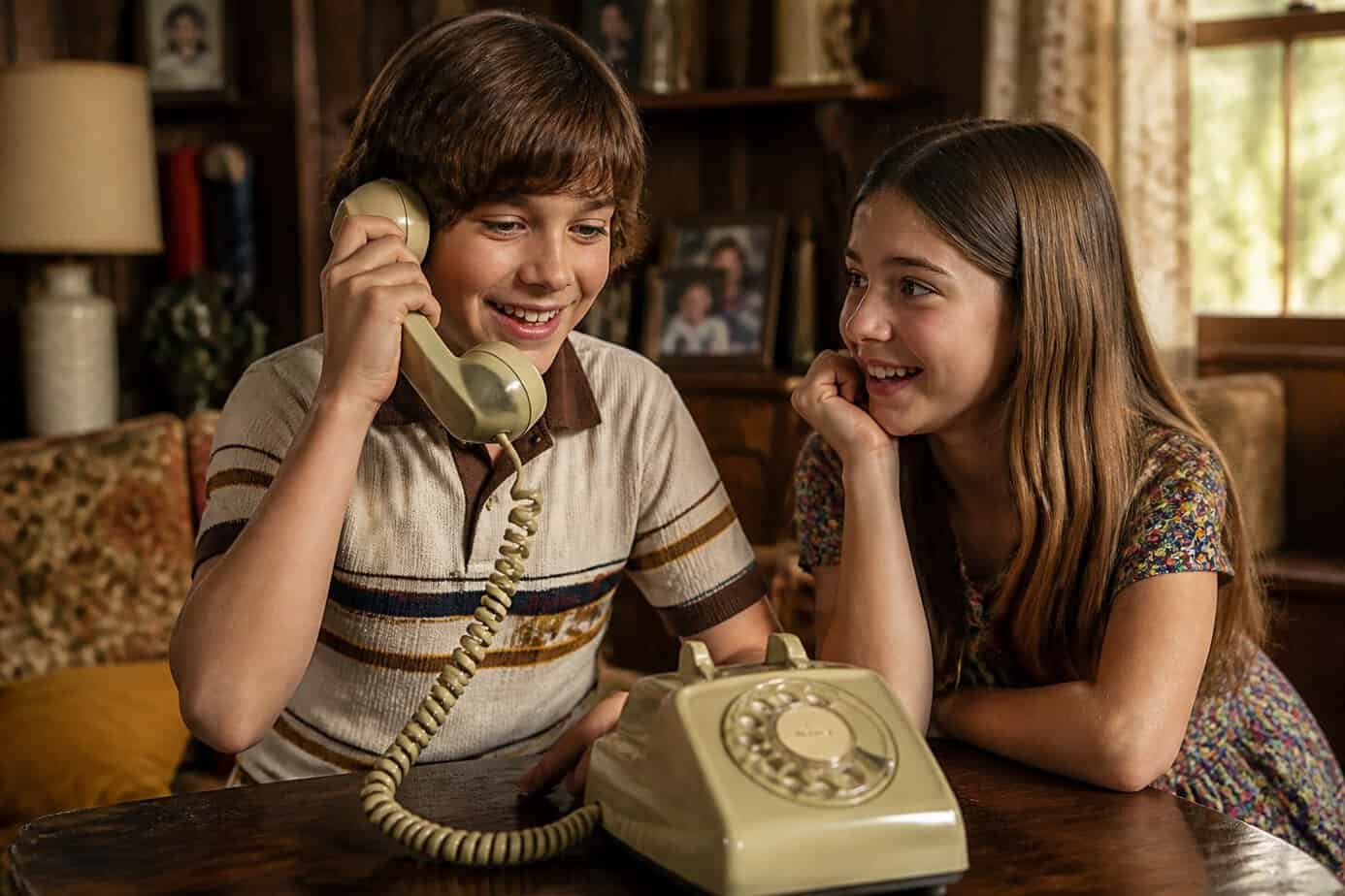 Two children in a 1970s home share a rotary telephone while calling friends.