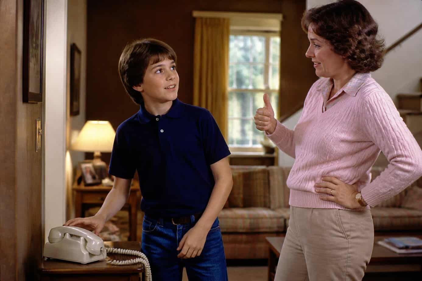 A child waits for a parent’s permission beside a corded home telephone.