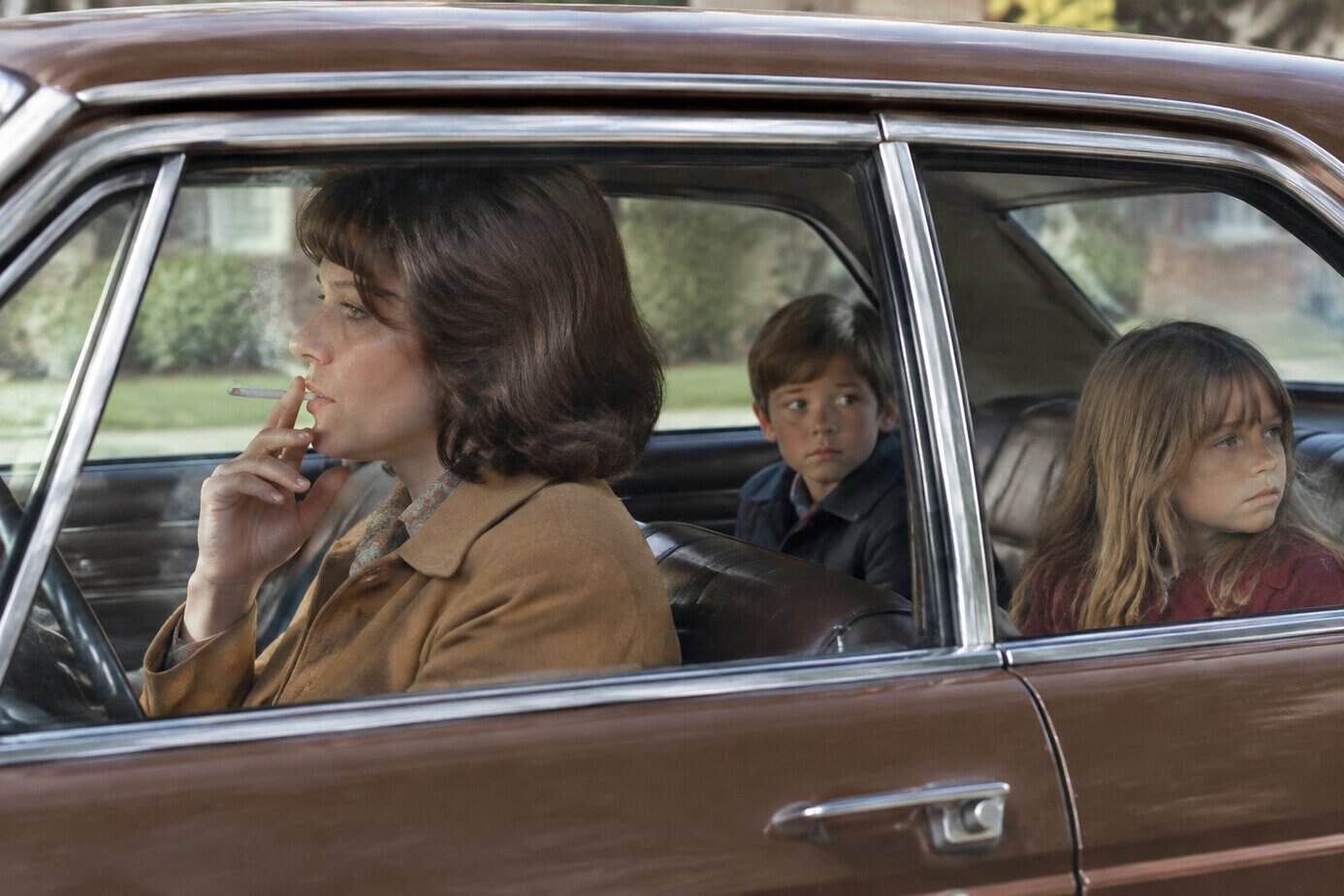 A mother smokes in the front seat of a car while two children sit in the back.