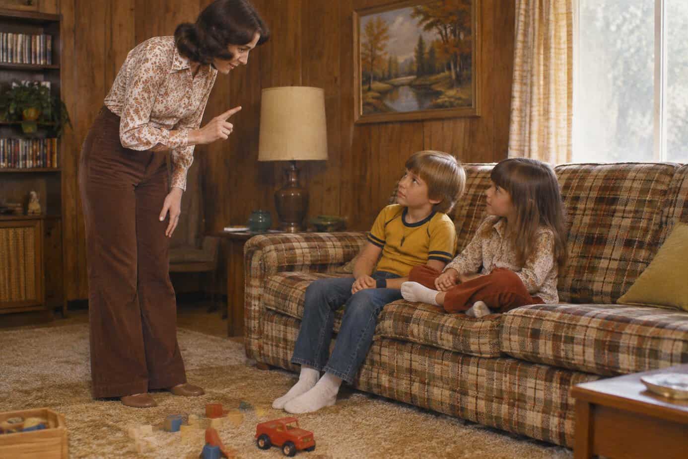 A 1970s mother wags her finger at two children sitting on a plaid sofa.