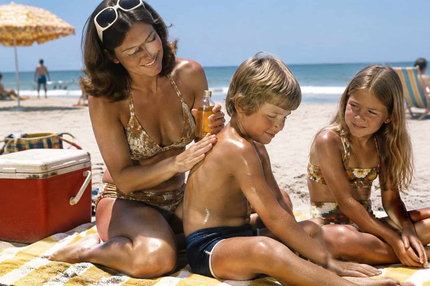 A mother applies baby oil to a child at a sunny beach while another child sits nearby.