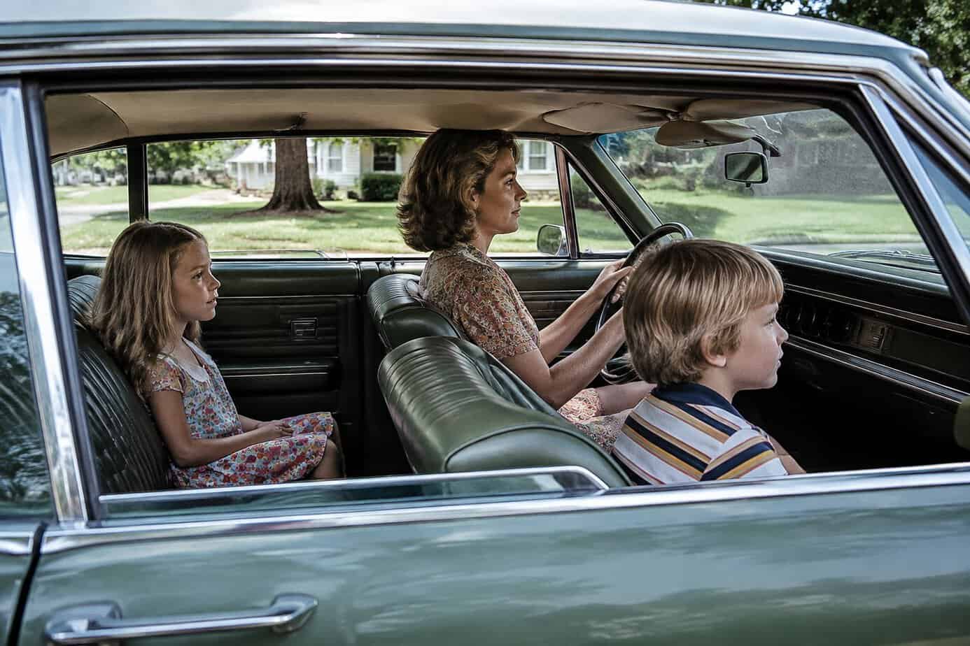 A mother drives with one child in the front seat and another in the back, neither using a seat belt.