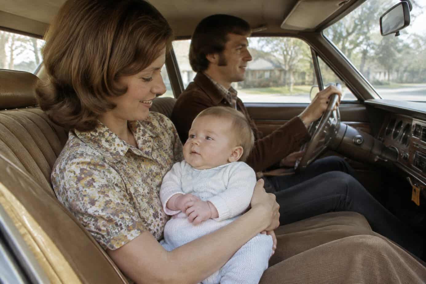 A mother holds her baby on her lap in the front passenger seat of a car.