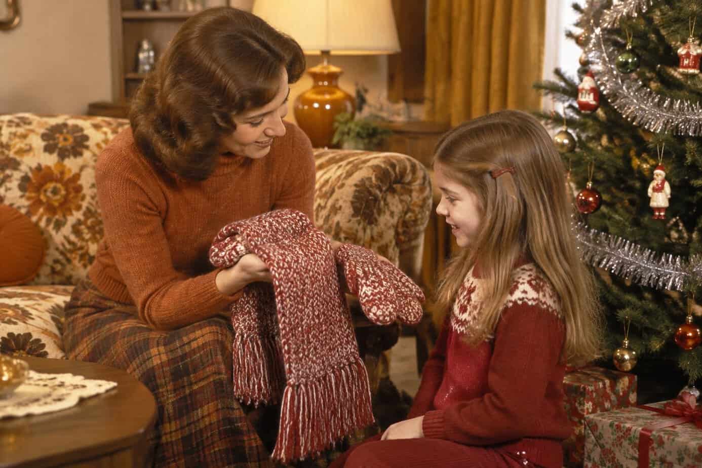 A mother gives her child a knitted scarf and mittens beside a Christmas tree.