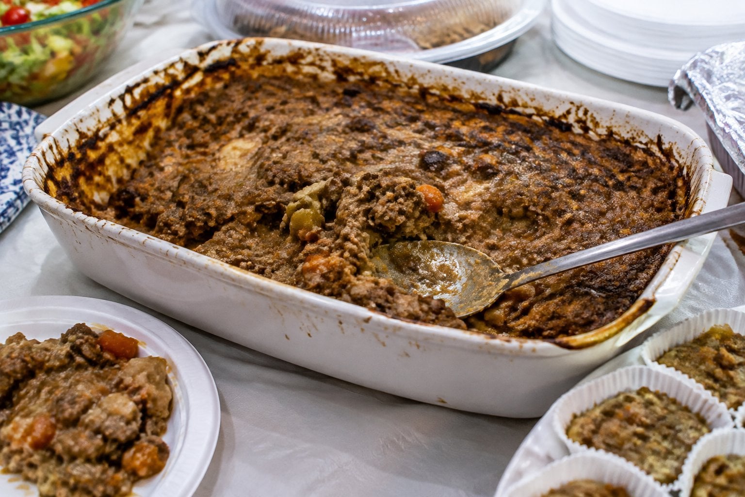 A casserole dish of lumpy meat and vegetables sits mostly untouched on a potluck buffet table.
