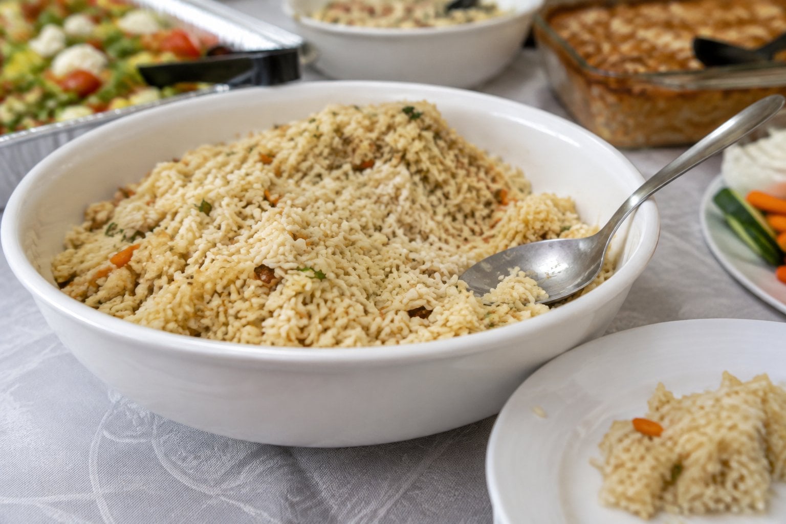 A bowl of dry-looking rice pilaf sits on a potluck buffet beside a serving spoon.