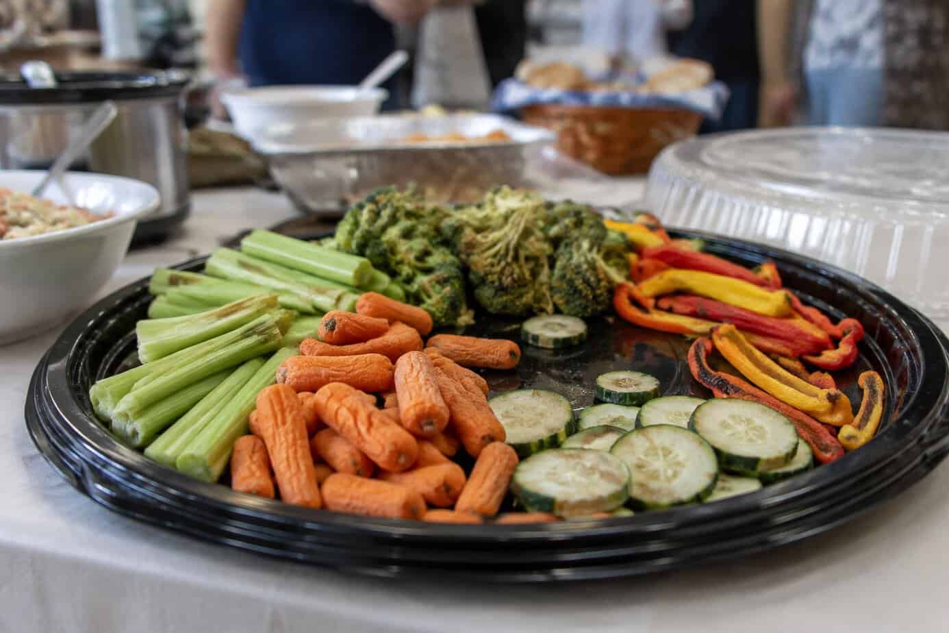 A tray of wilted, dried-out vegetables sits on a potluck buffet table.